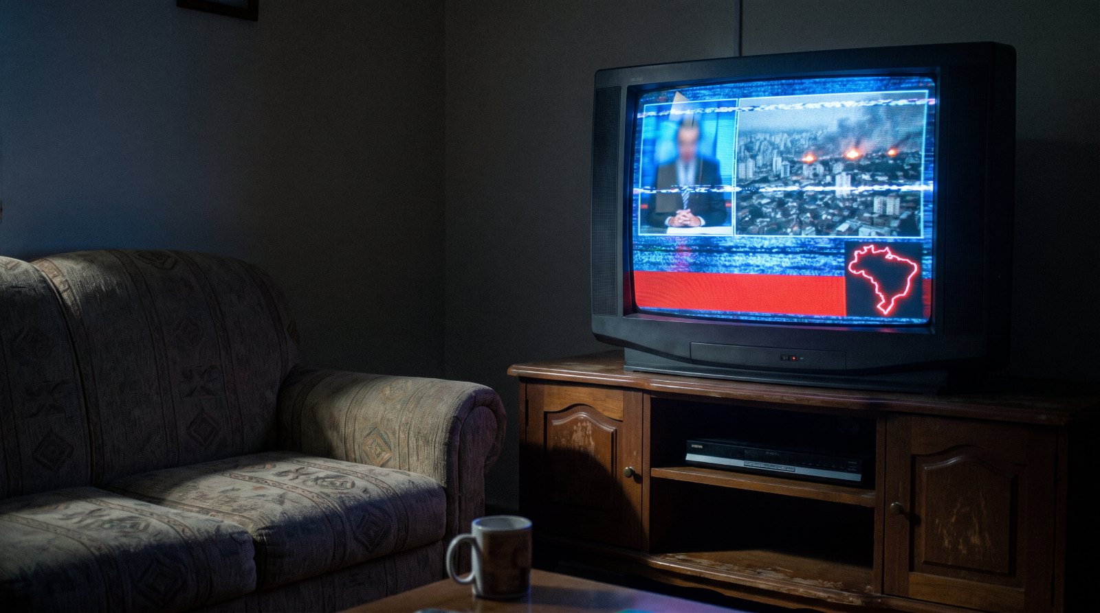 Uma TV velha numa sala escura mostrando um noticiário genérico com uma cidade em chamas